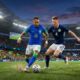 Sebuah foto aksi dinamis di dalam stadion yang penuh sesak pada malam hari, menampilkan pemain Brasil nomor 10 yang lincah mendribbel bola, sementara pemain Skotlandia nomor 4 berbadan tegap berusaha menghentikannya. Penonton di belakang mereka mengibarkan bendera Brasil dan Skotlandia.