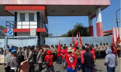 Massa pekerja alih daya Pertamina Balongan berseragam merah KASBI membawa bendera saat berunjuk rasa di depan gerbang fasilitas yang dijaga ketat aparat kepolisian.