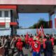 Massa pekerja alih daya Pertamina Balongan berseragam merah KASBI membawa bendera saat berunjuk rasa di depan gerbang fasilitas yang dijaga ketat aparat kepolisian.