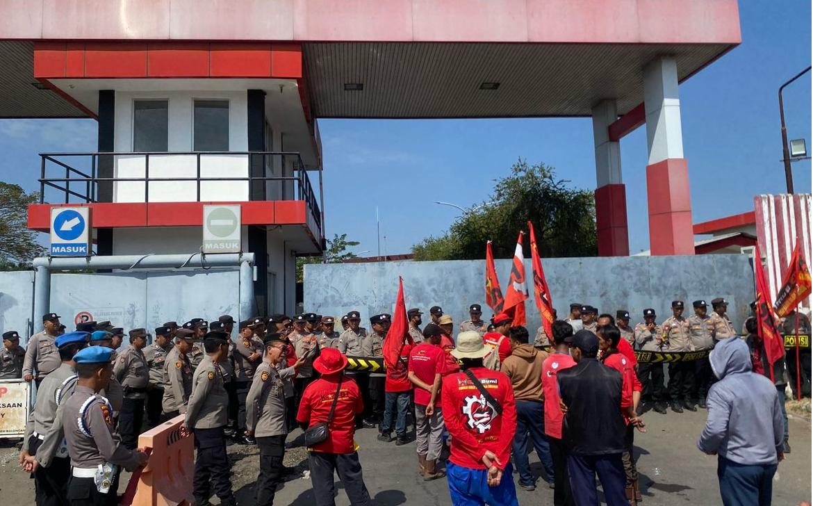 Massa pekerja alih daya Pertamina Balongan berseragam merah KASBI membawa bendera saat berunjuk rasa di depan gerbang fasilitas yang dijaga ketat aparat kepolisian.